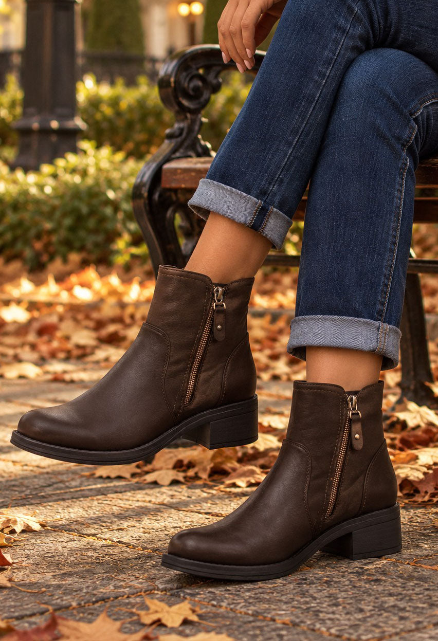 A person wearing dark brown leather ankle boots with side zippers, paired with cuffed blue jeans, sitting on a park bench with fallen autumn leaves on the ground.