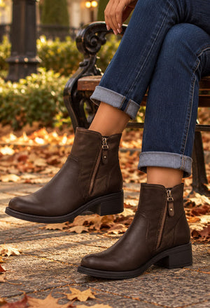 A person wearing dark brown leather ankle boots with side zippers, paired with cuffed blue jeans, sitting on a park bench with fallen autumn leaves on the ground.