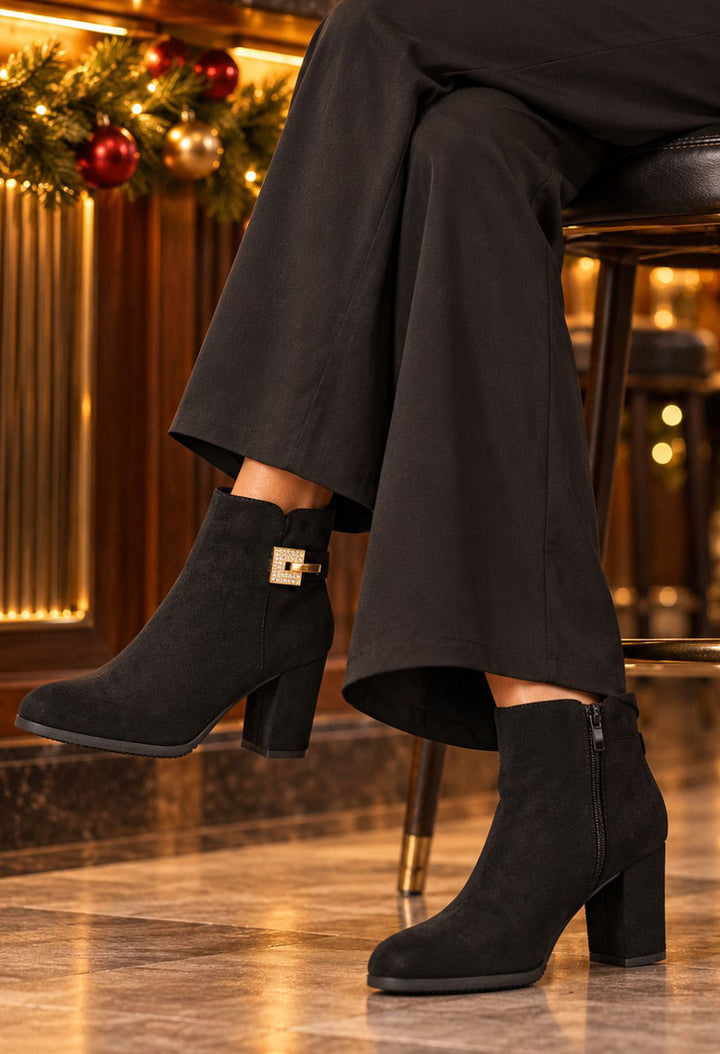 Black suede ankle boots with a block heel and a gold-tone decorative buckle on the side, shown on a model sitting on a stool in a warmly lit indoor setting.